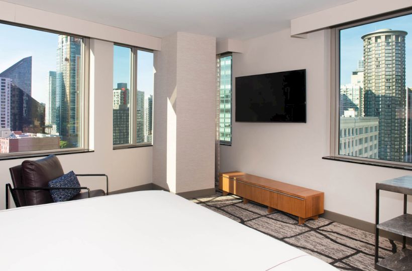 The image shows a modern hotel room with large windows offering city views, a wall-mounted TV, a chair, a low table, and a bed.