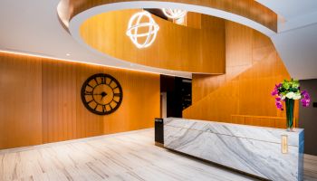 A modern reception area featuring a curved wooden staircase, a large wall clock, marble front desk, and a vase with flowers on the counter.