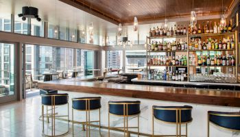 A modern bar with a sleek design features a long counter, high stools, and a fully stocked liquor shelf, with large windows offering city views.