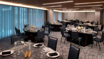The image shows a banquet hall with multiple round tables set with plates, silverware, and glasses. The room features dim lighting and elegant decor.