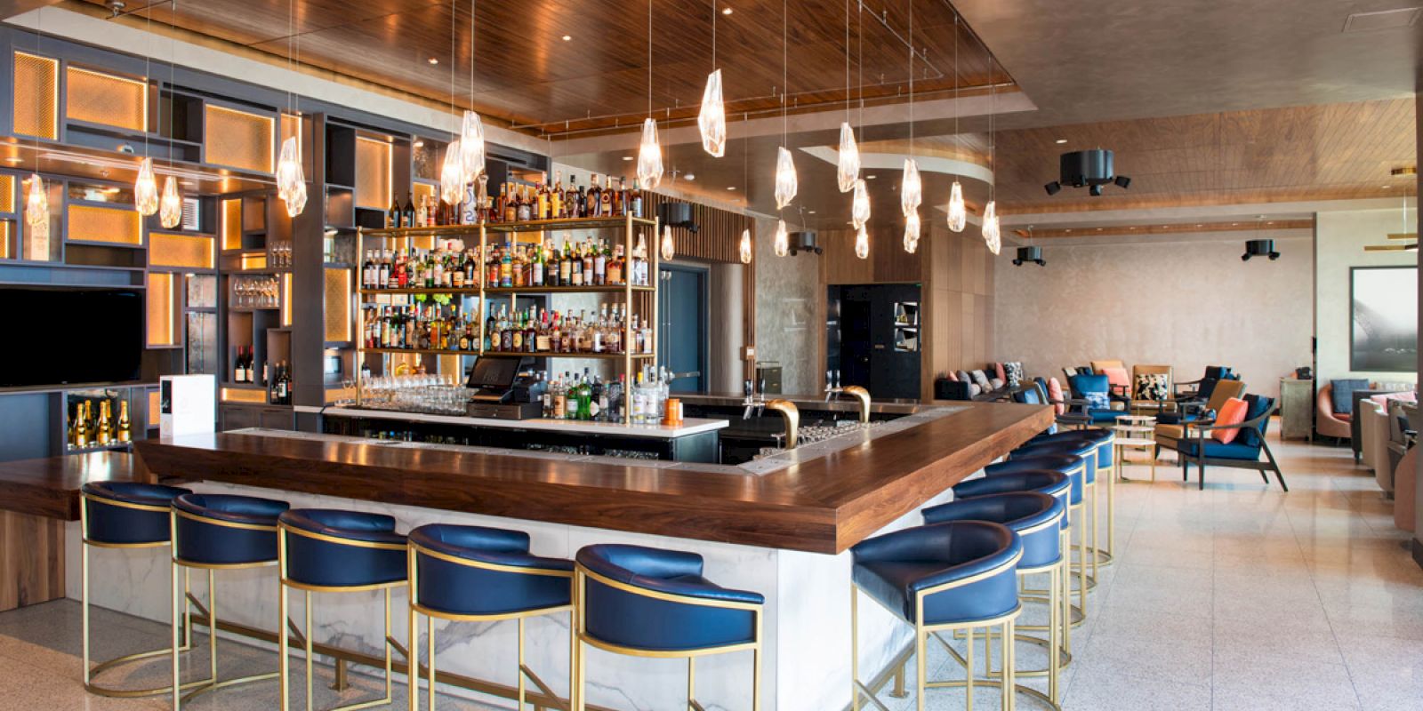 The image shows a modern bar with stylish seating, a well-stocked liquor shelf, pendant lighting, and a seating area in the background.