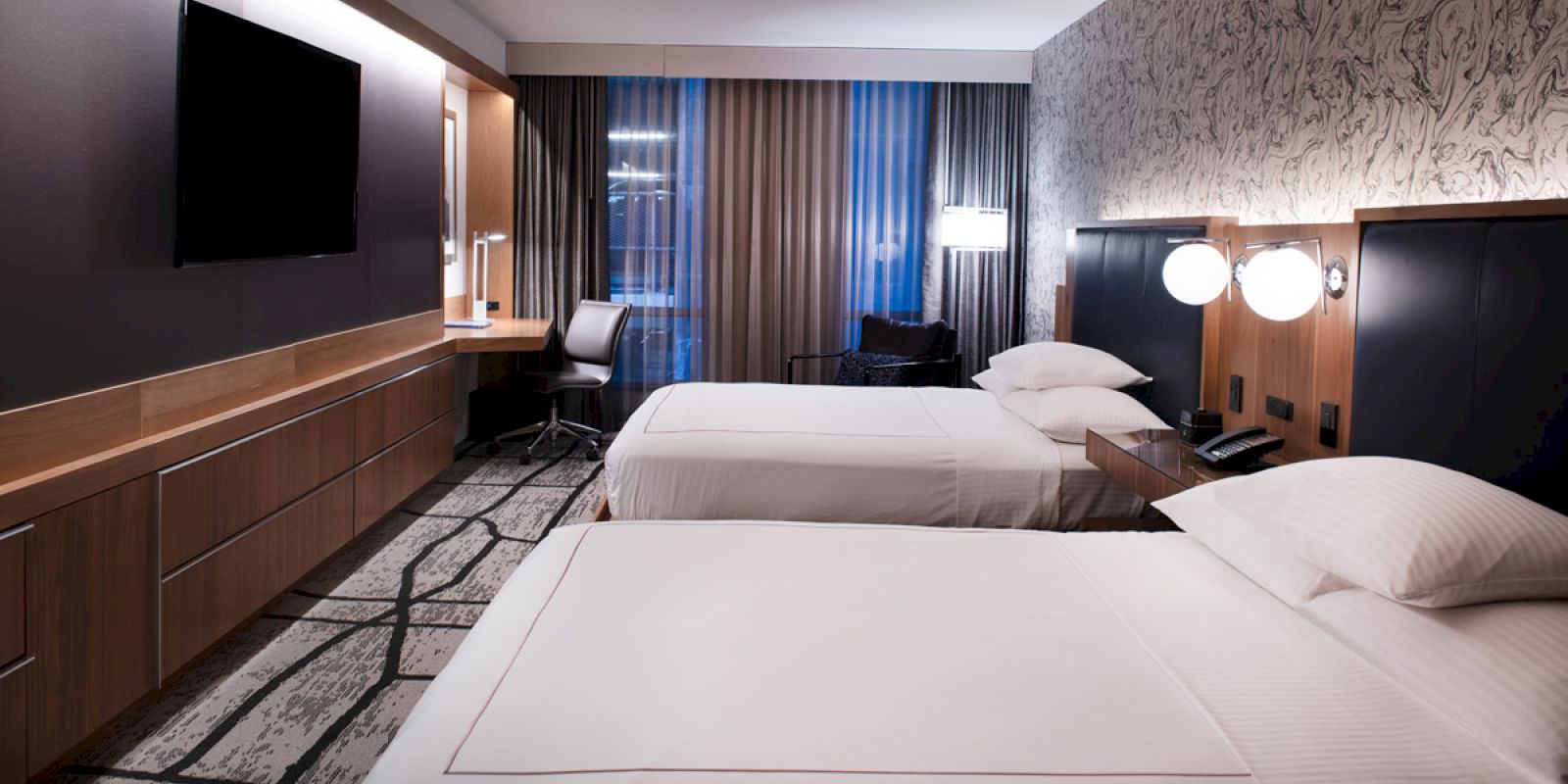 The image shows a modern hotel room with two neatly made twin beds, a large flat-screen TV, a desk with a chair, and a window with curtains.