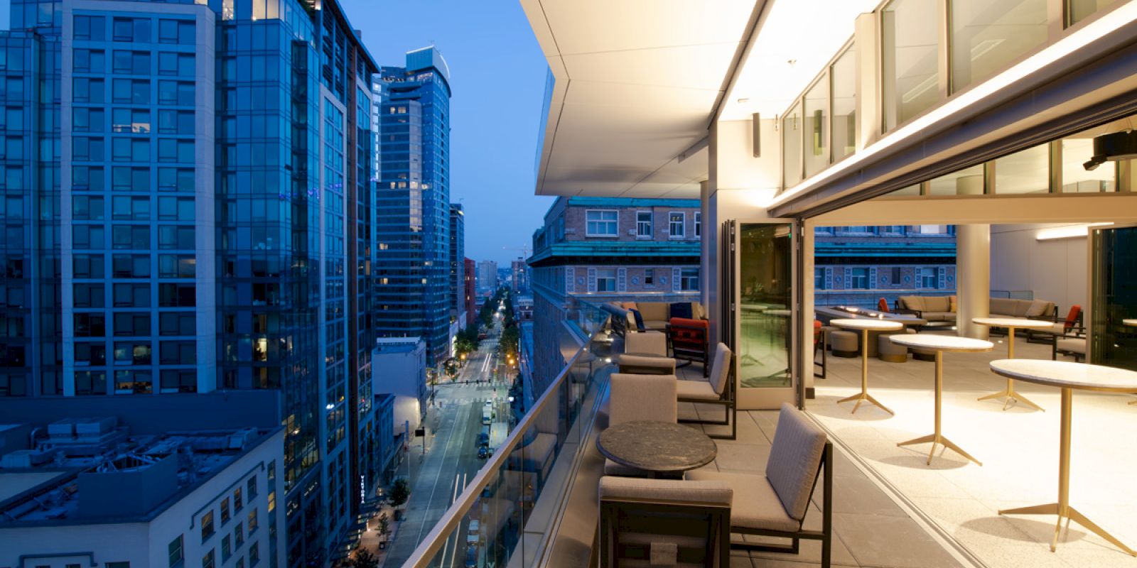 Modern cityscape from a high balcony at dusk, showing tall buildings and a stylish, illuminated outdoor seating area.