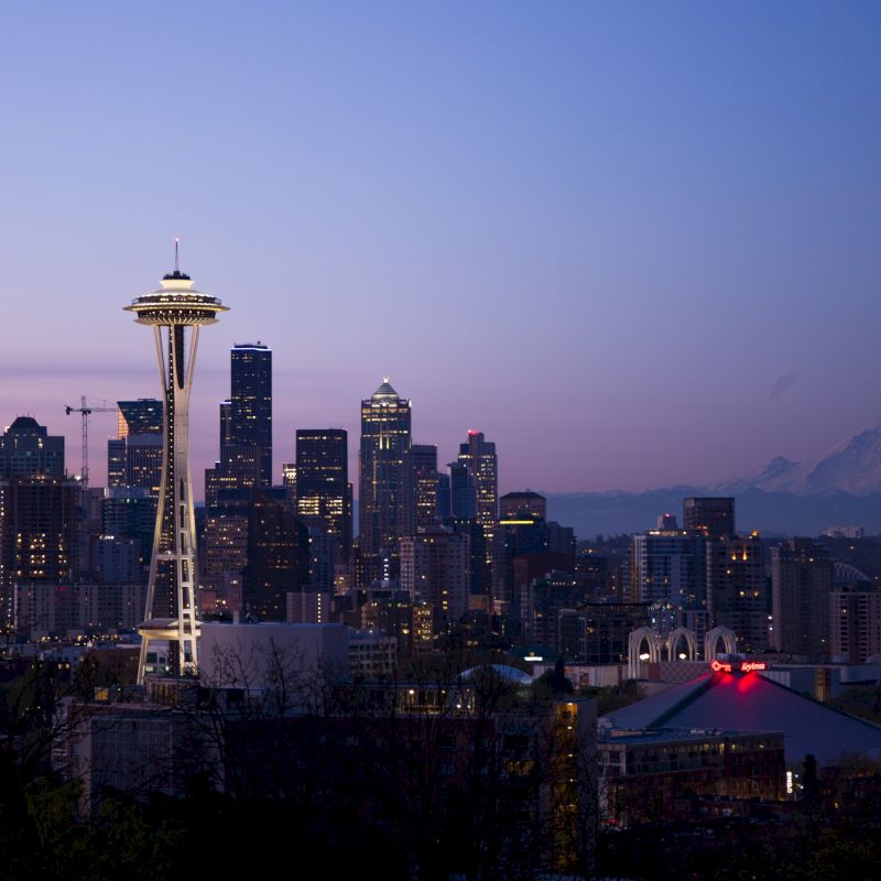 The image shows a city skyline at dusk, featuring the Space Needle, numerous skyscrapers, and a mountain in the background under a twilight sky.