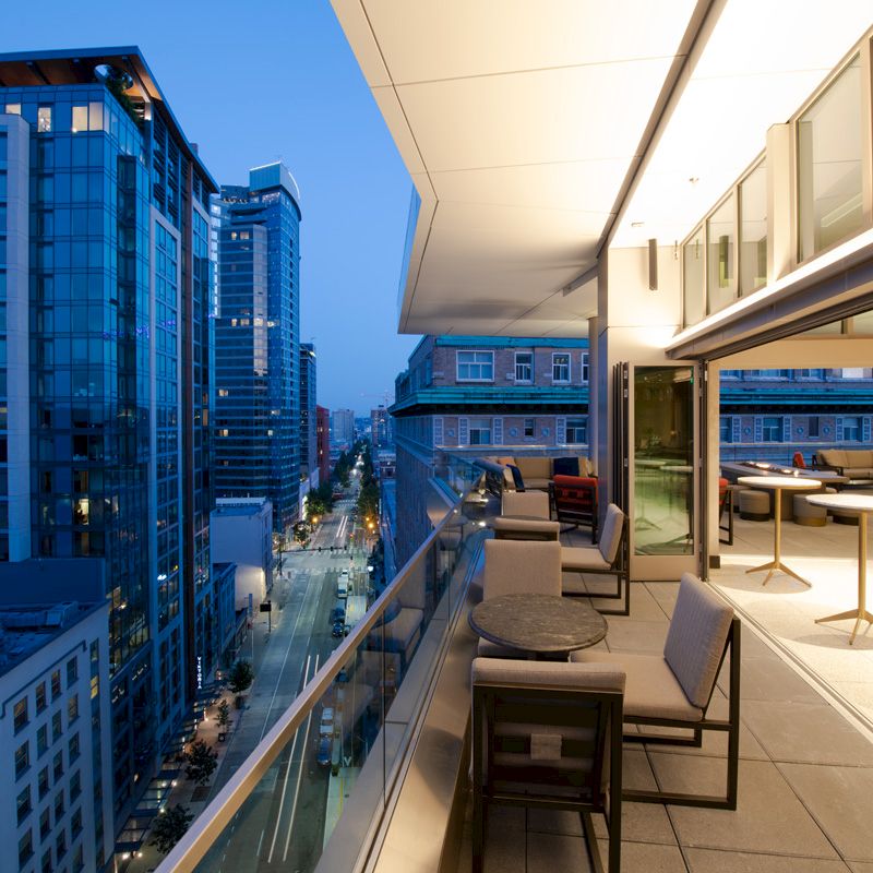 A modern urban balcony view at dusk features seating and tables, with a cityscape of tall buildings and streetlights in the background.