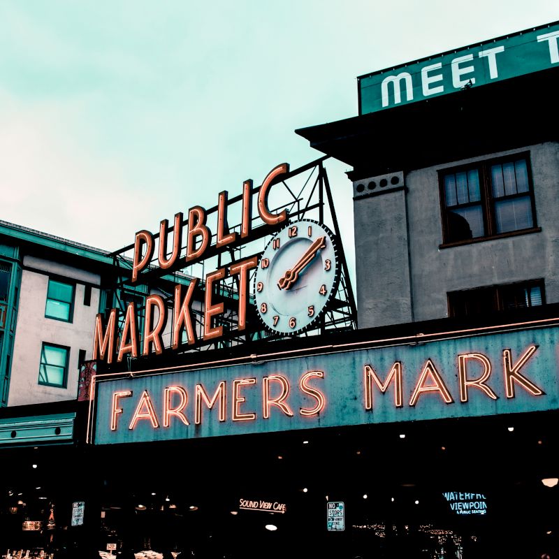 The image shows an iconic outdoor market scene with a large sign reading "Public Market" and "Farmers Market" with a clock, and buildings in the background.