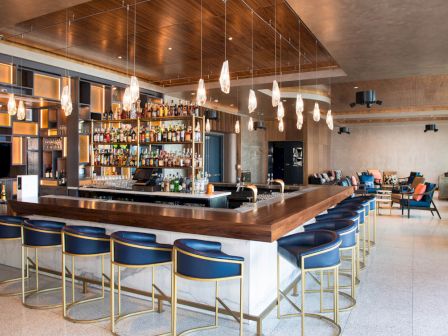 A modern bar with a large selection of bottles, stylish seating, hanging lights, and a cozy seating area in the background is shown in the image.