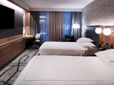 A modern hotel room with two neatly made beds, a large TV on the wall, a desk with a chair, and contemporary decor and lighting.