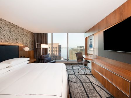 The image depicts a modern hotel room with a large bed, a desk with a chair, a flat-screen TV, and a view of buildings and water outside.