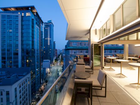 The image shows a modern urban balcony with seating, overlooking a city street lined with tall buildings at dusk.