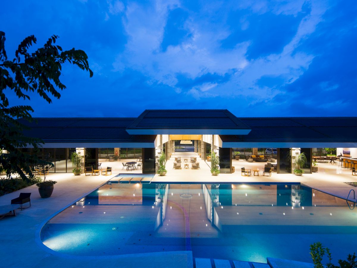 A lit swimming pool in front of a modern, open-air building with lounge areas, surrounded by trees under a twilight sky.
