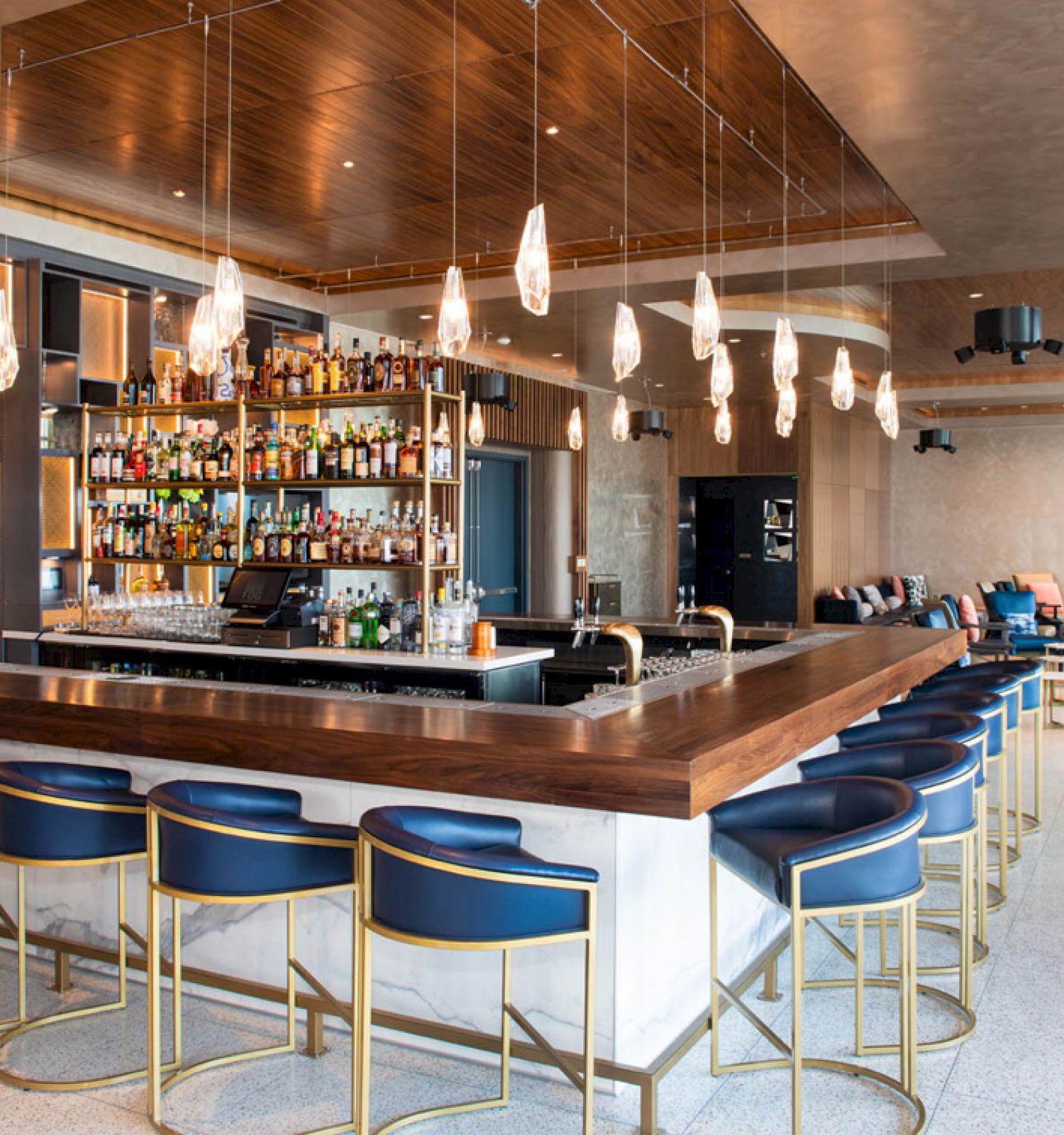 The image shows a modern bar with a counter surrounded by blue and gold stools, shelves stocked with bottles, and contemporary light fixtures.