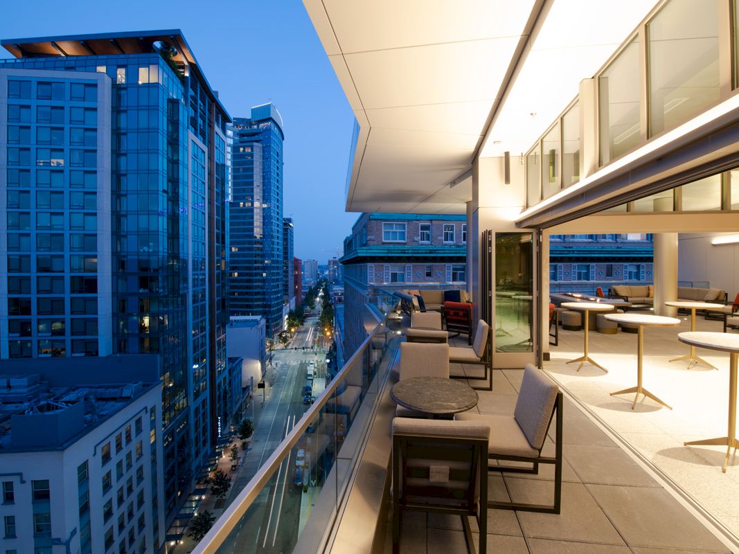 A modern cityscape at dusk viewed from a balcony with tables, chairs, and lounge spaces, lined by tall buildings and a busy street below.