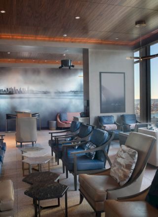 A modern, stylish lounge with comfortable seating, large windows offering a cityscape view, a mural on the wall, and various elegant tables.