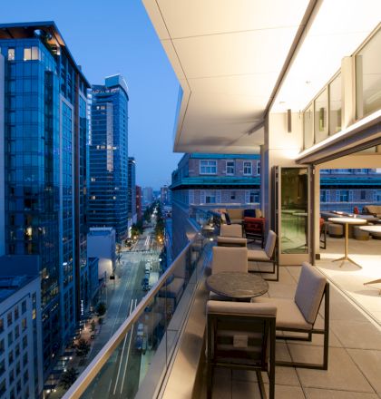 A modern urban balcony with seating and tables overlooks a city street lined with tall glass buildings at dusk, with lights illuminating softly.
