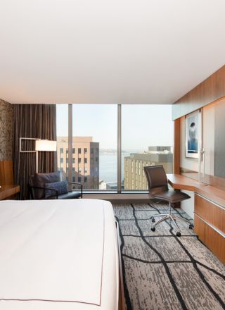 A modern hotel room with a large bed, desk, chair, flat-screen TV, and cityscape window view. The room features contemporary decor and furnishings.