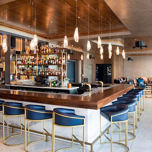 The image shows a modern bar with a wooden counter, blue barstools, shelves of liquor bottles, stylish pendant lights, and a lounge area in the background.