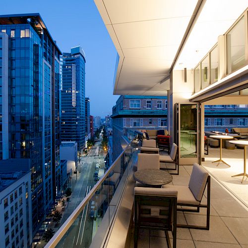 A modern cityscape with tall buildings and a spacious balcony featuring tables and chairs, overlooking a busy street at dusk.
