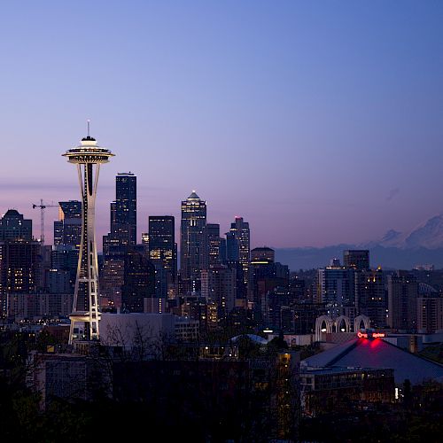 The image shows a skyline view of Seattle at dusk, featuring the Space Needle prominently, with Mount Rainier visible in the background.