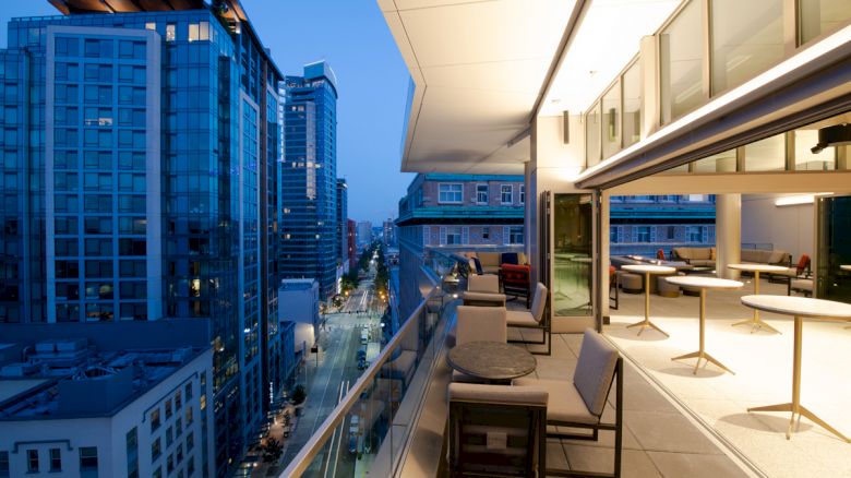 A modern balcony with seating overlooks a city's skyscrapers at twilight. The interior space features tables and large glass doors for a seamless flow.