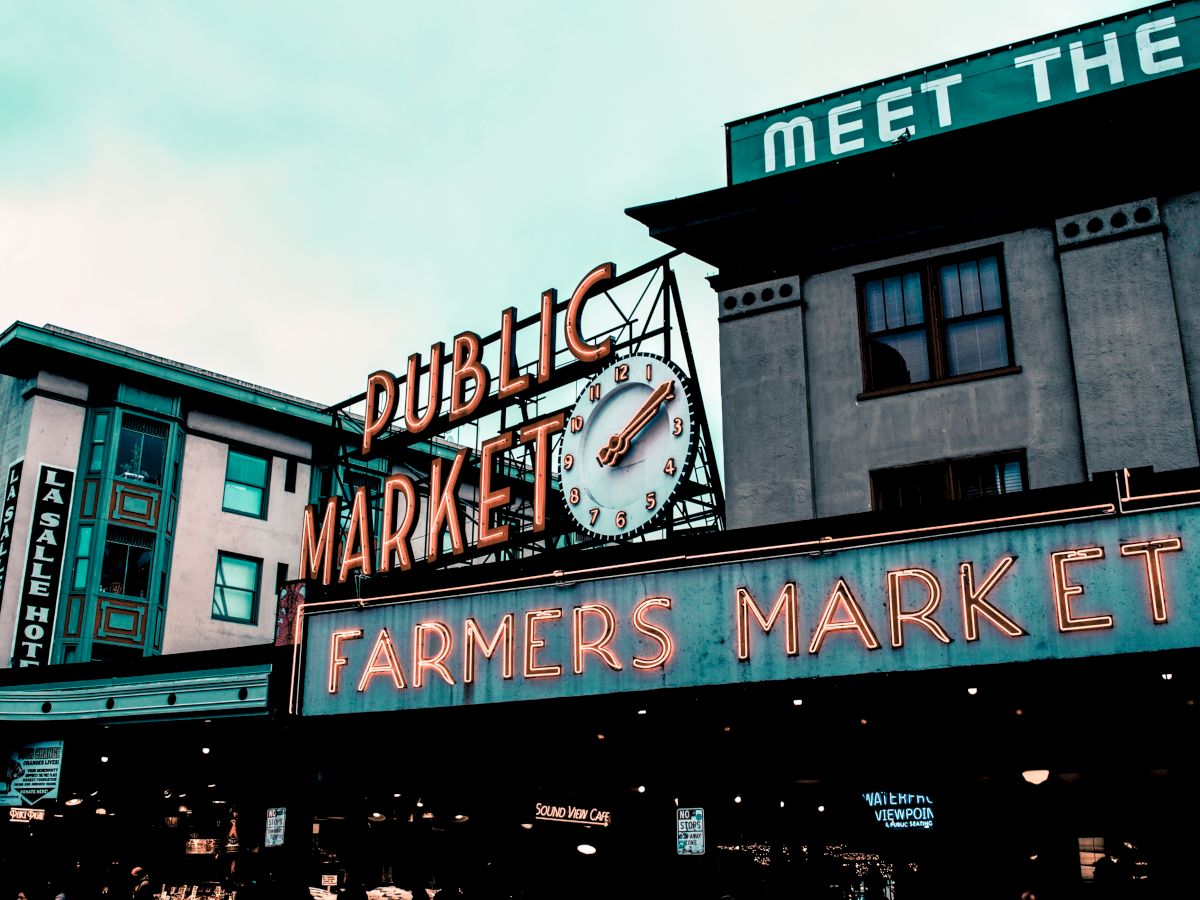 The image shows the iconic neon signs for "Public Market" and "Farmers Market" at Pike Place Market in Seattle, with surrounding building structures.