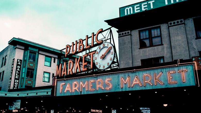 The image shows the iconic neon signs for "Public Market" and "Farmers Market" at Pike Place Market in Seattle, with surrounding building structures.