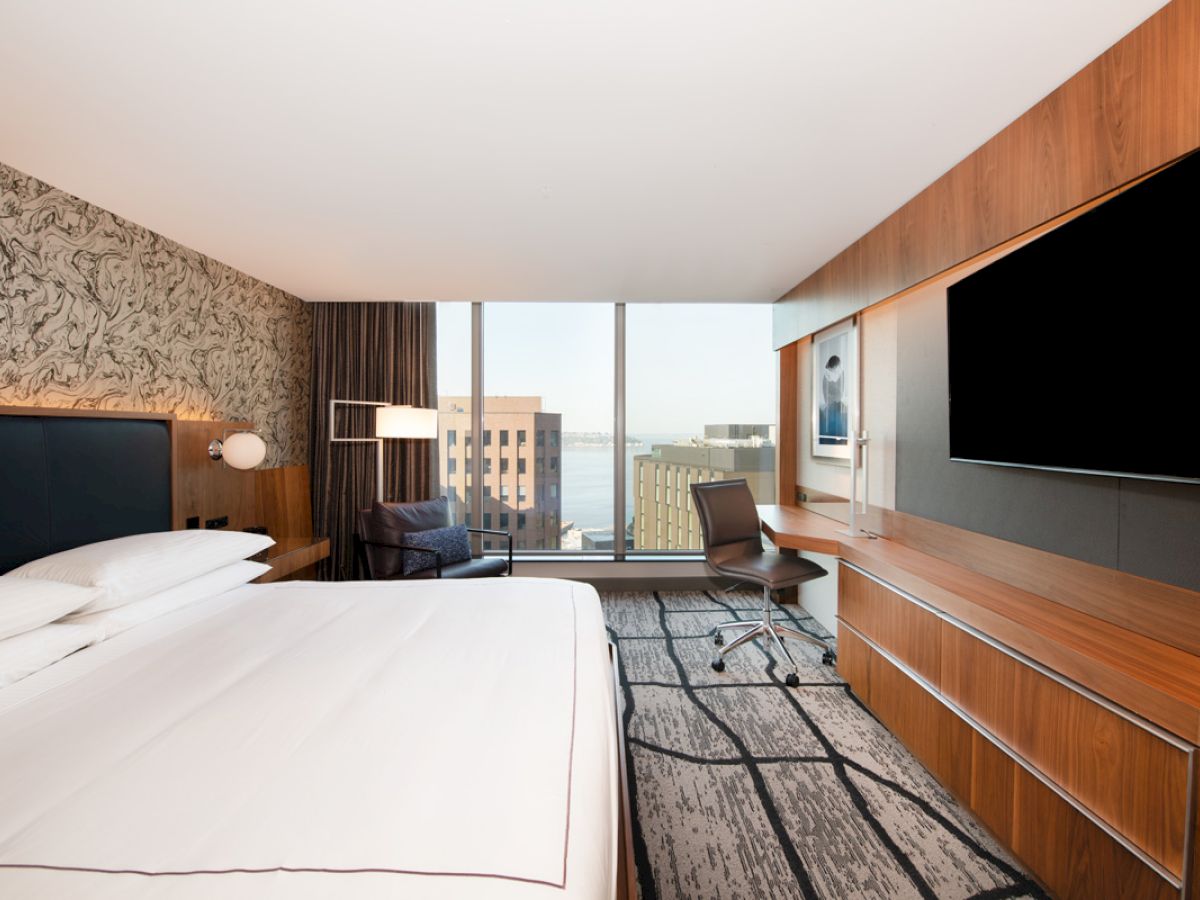 A modern hotel room features a large bed, wall-mounted TV, desk with chair, and city view through large windows.