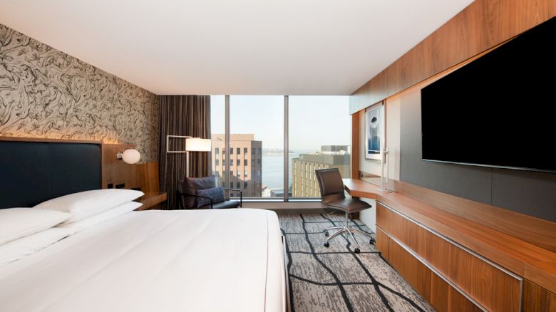 A modern hotel room features a large bed, wall-mounted TV, desk with chair, and city view through large windows.