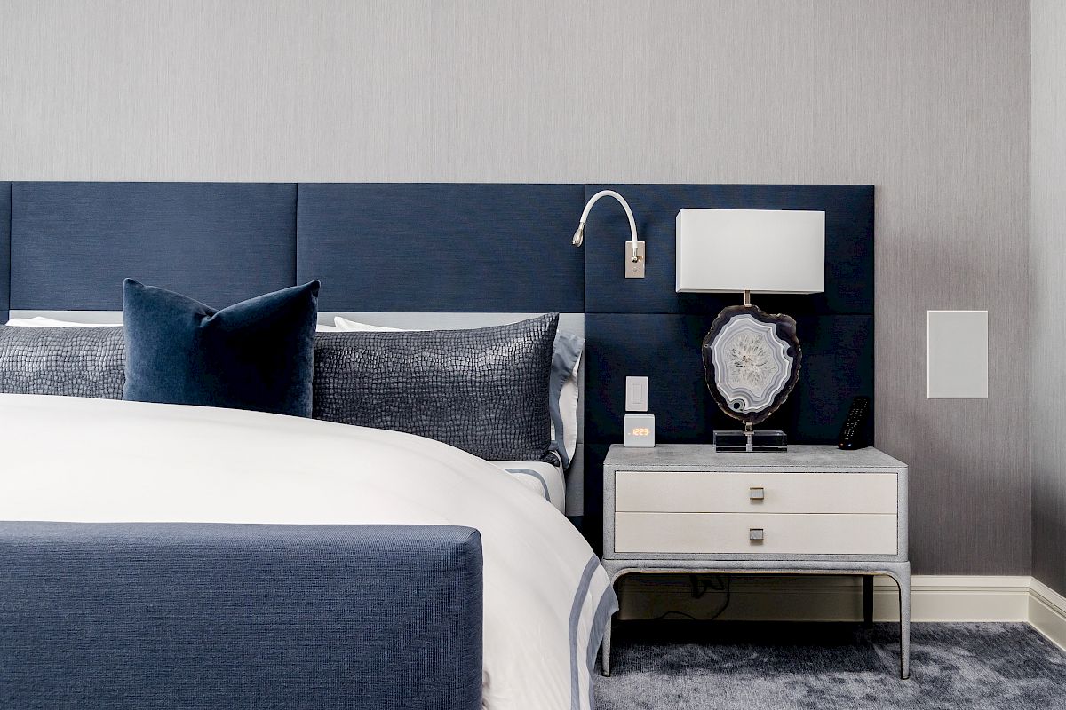 A modern bedroom features a blue and white color scheme with a bed, upholstered headboard, nightstand, lamp, and decorative items.