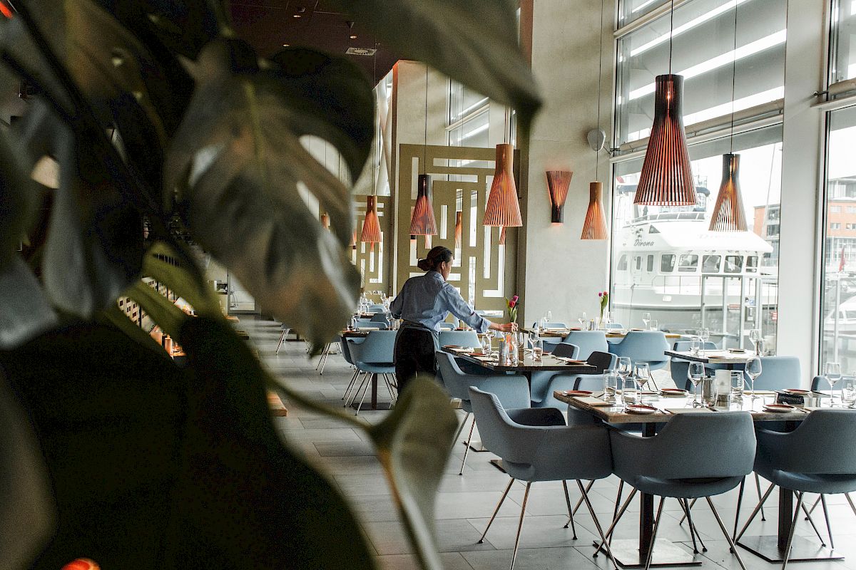 A restaurant with modern decor, where a person is setting tables near large windows with ample natural light.