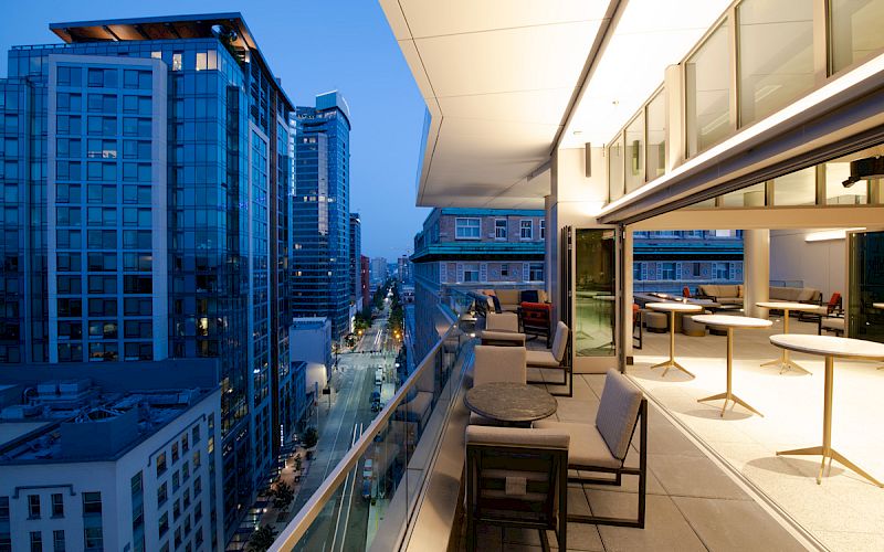 A modern urban balcony with seating overlooks a cityscape with tall buildings at dusk; indoor seating is also visible through glass doors.