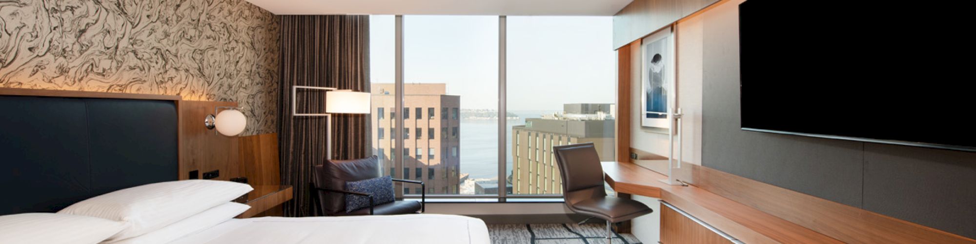 A modern hotel room features a large bed, desk with chair, armchair by the window, flat-screen TV, and a view of buildings and water.